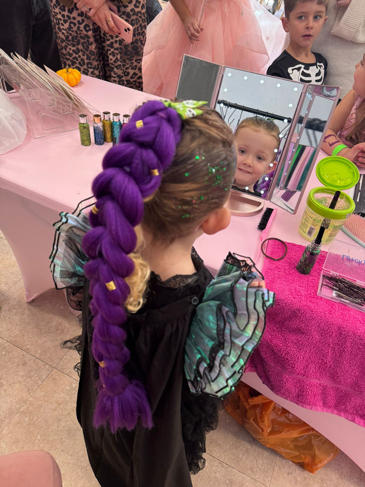 Child with purple braids in a salon setting with mirrors and products.