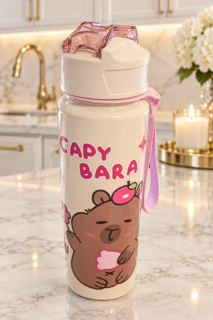 A white insulated water bottle with a cartoon bear graphic and the text 'CAPY BARA' in pink, shown on a marble kitchen counter with a candle and flowers in the background.